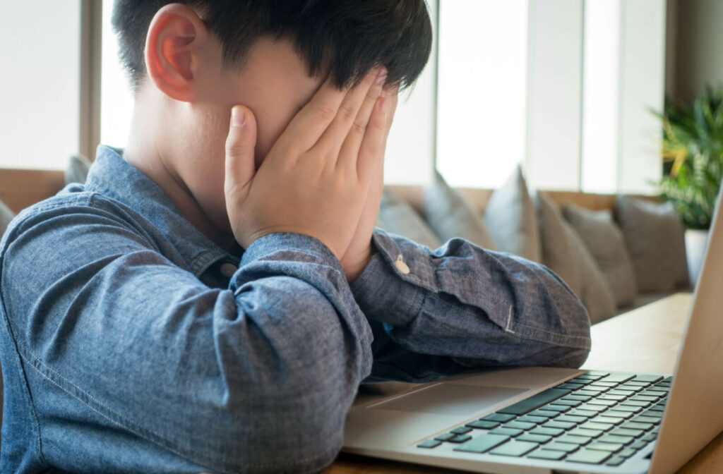 Having blurry vision, which is a sign of digital eye strain, a young boy using a laptop covers his face with his hands.