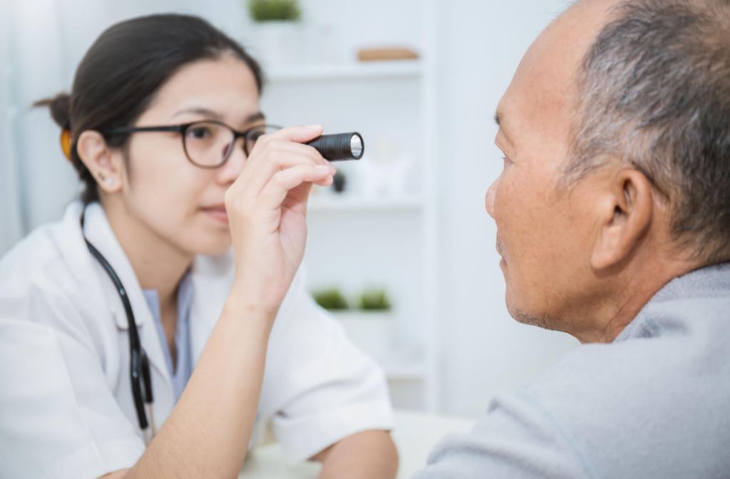 A senior man facing his optometrist as she shines a small flashlight into his eye.