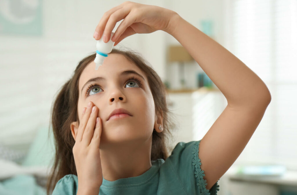 A child holding a small bottle of eye drops in her left hand and putting them on her right eye while she uses her left fingers to pull her eyelid down.