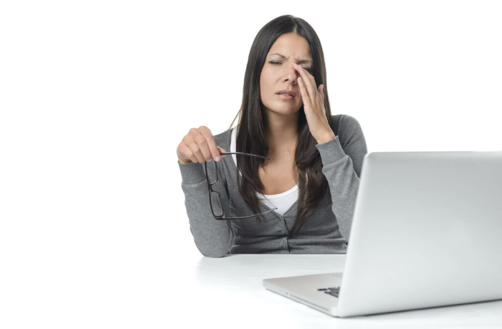 A woman sitting at a desk with a laptop as she holds her glasses in her right and and rubs her eye.