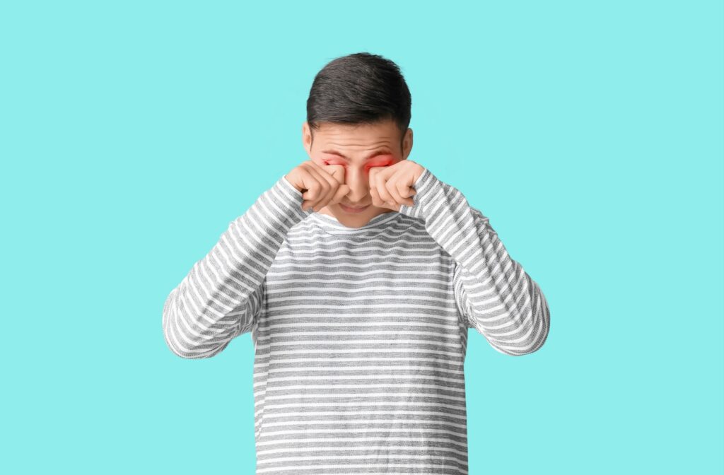  A person wearing a striped shirtstands in front of a blue background. They are rubbing their dry, red, irritated eyes.