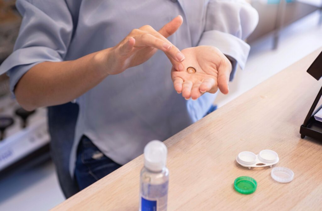 Contact lens, contact solution bottle, and lens case neatly arranged on a table with a person’s hand holding a lens.