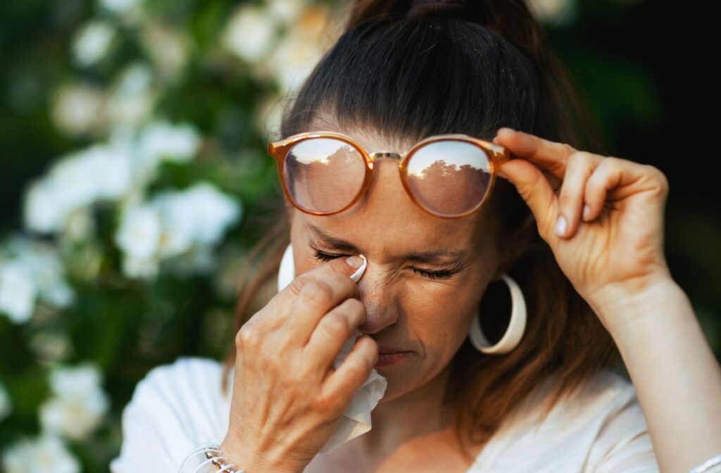 A person outside, lifting their glasses up to wipe their itchy eyes from allergies with a tissue.