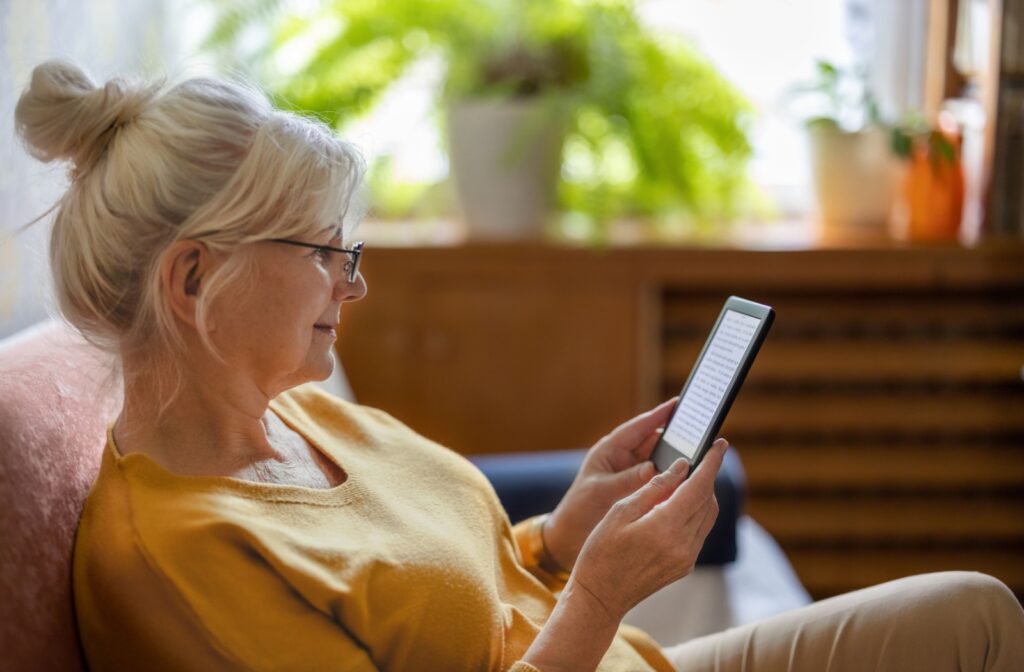A person sitting on the couch, reading a e-book on their kindle, wearing glasses to prevent eye strain.