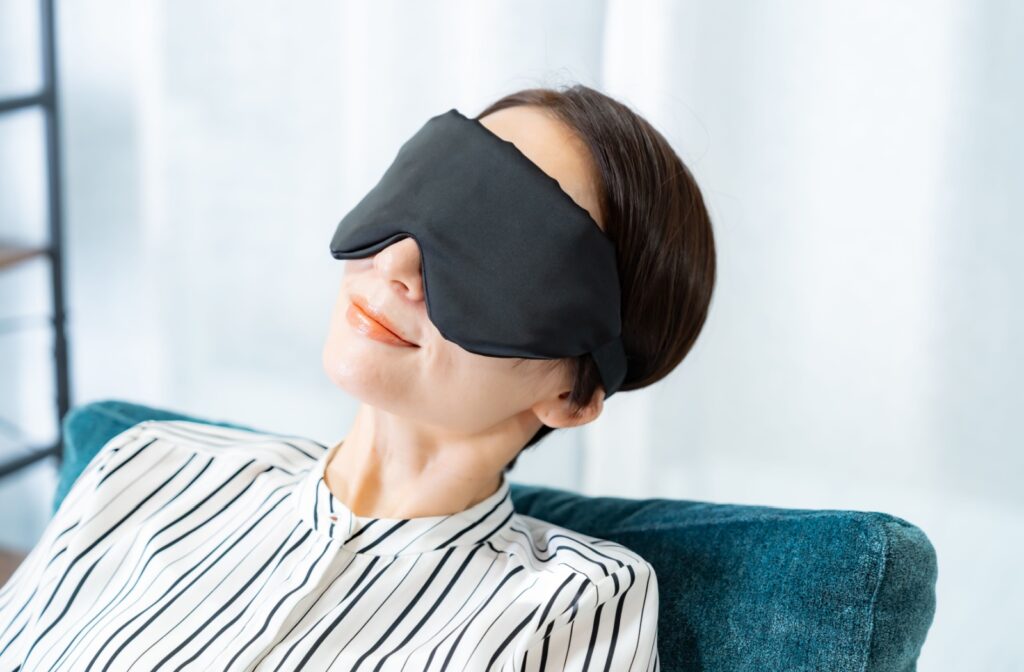 A person sitting on a couch, wearing a warming eye mask to help relieve symptoms of dry eye and eye strain.